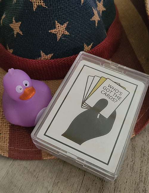 Card game packaging with 'Who's Got the Cards?' text and a purple rubber duck on a checkered fabric background.